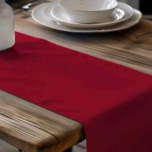 May include: A deep red table runner on a wooden table, with white ceramic plates, bowls, and a vase with dried flowers. The runner adds a pop of color to the neutral tones of the table setting.