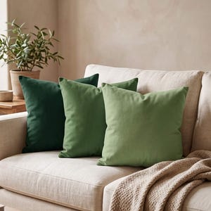 May include: Three square throw pillows arranged in a row on a sofa. The pillows feature a solid color design in a gradient of green shades, ranging from a deep forest green on the left to a medium olive green and a lighter sage green on the right.