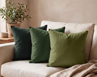 May include: Three square throw pillows arranged in a row on a light beige sofa. The pillows feature a smooth, solid-colored fabric in a gradient of green shades, ranging from a deep forest green on the left to a lighter olive green on the right.