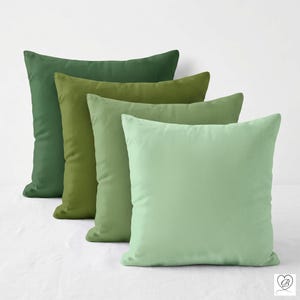 May include: A set of four square throw pillows arranged in a cascading row, each featuring a different shade of green. The colors transition from a deep forest green in the back to a light, soft mint green in the front, with smooth fabric.