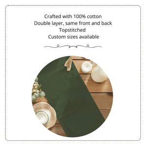 以下が含まれることがあります： 100%コットン製のダークオリーブグリーンのテーブルランナー。テキストには「Crafted with 100% cotton, Double layer, same front and back, Topstitched, Custom sizes available」と記載されています。木製の表面に白いキャンドルと共に配置されています。