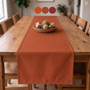 May include: A solid burnt orange rectangular table runner made of fabric, centered on a wooden dining table. A wooden bowl sits on the runner, filled with decorative spheres made of woven twine and natural dried plant elements.
