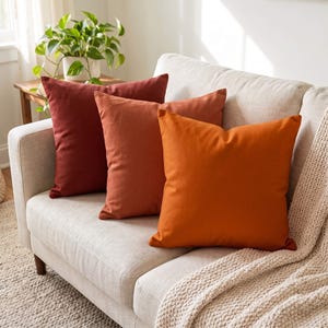May include: Three square throw pillows in a gradient of warm colors, ranging from a deep dark red to a medium terracotta and a bright orange, arranged in a row on a light-colored sofa with a textured cream-colored throw blanket draped over the side.