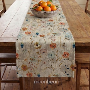 May include: A long rectangular table runner featuring a botanical print with orange, yellow, and blue flowers on a light beige background. A gray bowl filled with several bright orange oranges sits centered on the runner, which is laid flat.