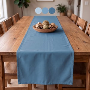 May include: A solid blue rectangular table runner made of fabric, centered on a wooden dining table. A wooden bowl filled with decorative natural fiber spheres and dried botanical elements sits on top of the runner.
