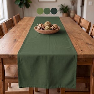 May include: A solid olive green rectangular table runner made of fabric with hemmed edges, centered on a wooden dining table. A wooden bowl containing decorative spheres and dried natural elements sits on top of the runner.