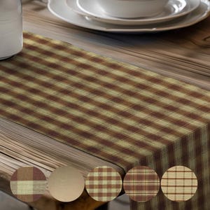 May include: A brown and tan plaid table runner. The runner is rectangular and features a repeating pattern of intersecting lines, creating a checkered design. The fabric has a rustic, homespun appearance. The runner is displayed on a wooden table, with white dishes in the background.
