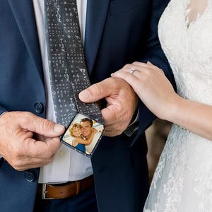 May include: A dark grey necktie featuring a repeating pattern of small white symbols and a rectangular photograph printed on the underside of the tip, showing a young child embracing an adult, worn with a navy blue suit jacket and white shirt.