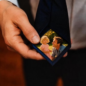 Peut inclure: Une cravate bleu marine foncé présentant une photographie personnalisée imprimée sur sa face intérieure. L'image visible montre un jeune enfant en robe rose debout à côté d'un adulte en chemise bleue, dans un cadre extérieur ensoleillé.