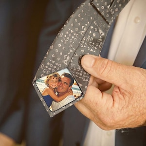 Puede incluir: Una corbata gris con un patrón geométrico repetitivo. La corbata presenta una fotografía enmarcada de un hombre y una niña pequeña abrazándose. La corbata es sostenida por una persona que lleva un traje.