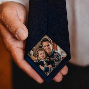 May include: A dark navy blue necktie featuring a triangular inset on the back panel. The inset displays a color photograph of an adult and a young child smiling together. The tie fabric has a subtle, fine-grained texture throughout the surface.