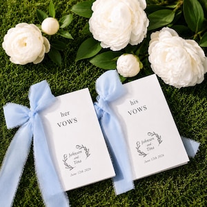 Può includere: Due libretti per i voti nuziali bianchi con nastri in chiffon azzurro. Ogni libretto presenta testo nero con scritto 'her VOWS' o 'his VOWS', un grafico a corona floreale, i nomi 'Johnson and Tina' e la data 'June 12th 2026' sulle copertine.