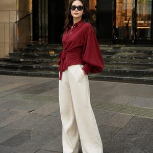 May include: A deep red long-sleeved button-down shirt with a tie-front waist and voluminous sleeves paired with high-waisted cream-colored wide-leg trousers and pointed-toe shoes, accessorized with black sunglasses.