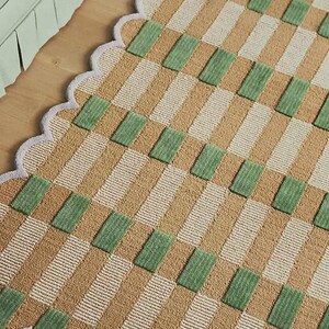 May include: A rectangular rug with a scalloped edge. The rug has a repeating pattern of tan, white, and green rectangles. The rug is made of a woven material and is placed on a wooden floor.