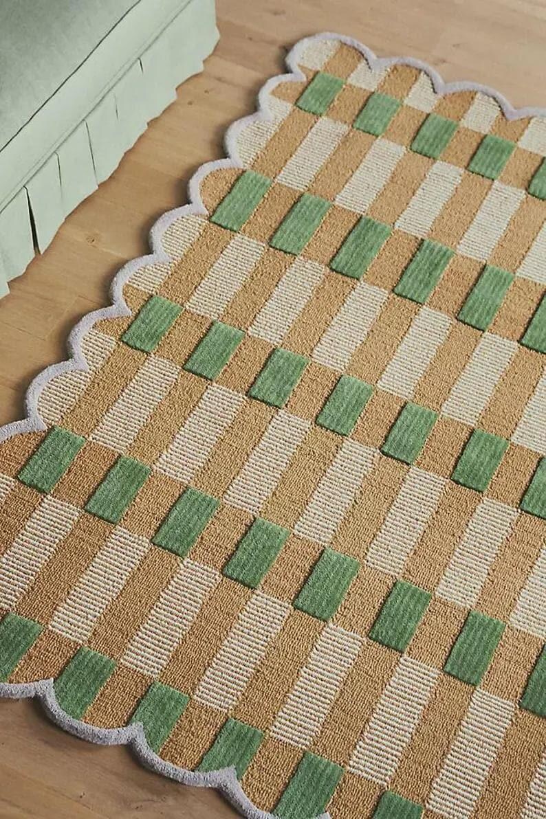 May include: A rectangular rug with a scalloped edge. The rug has a repeating pattern of tan, white, and green rectangles. The rug is made of a woven material and is placed on a wooden floor.