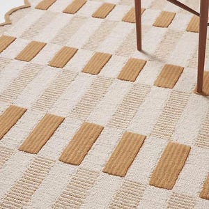 May include: A beige and white patterned rug with a scalloped edge. The rug features alternating rows of beige rectangular blocks and white striped sections. A wooden chair leg is visible.
