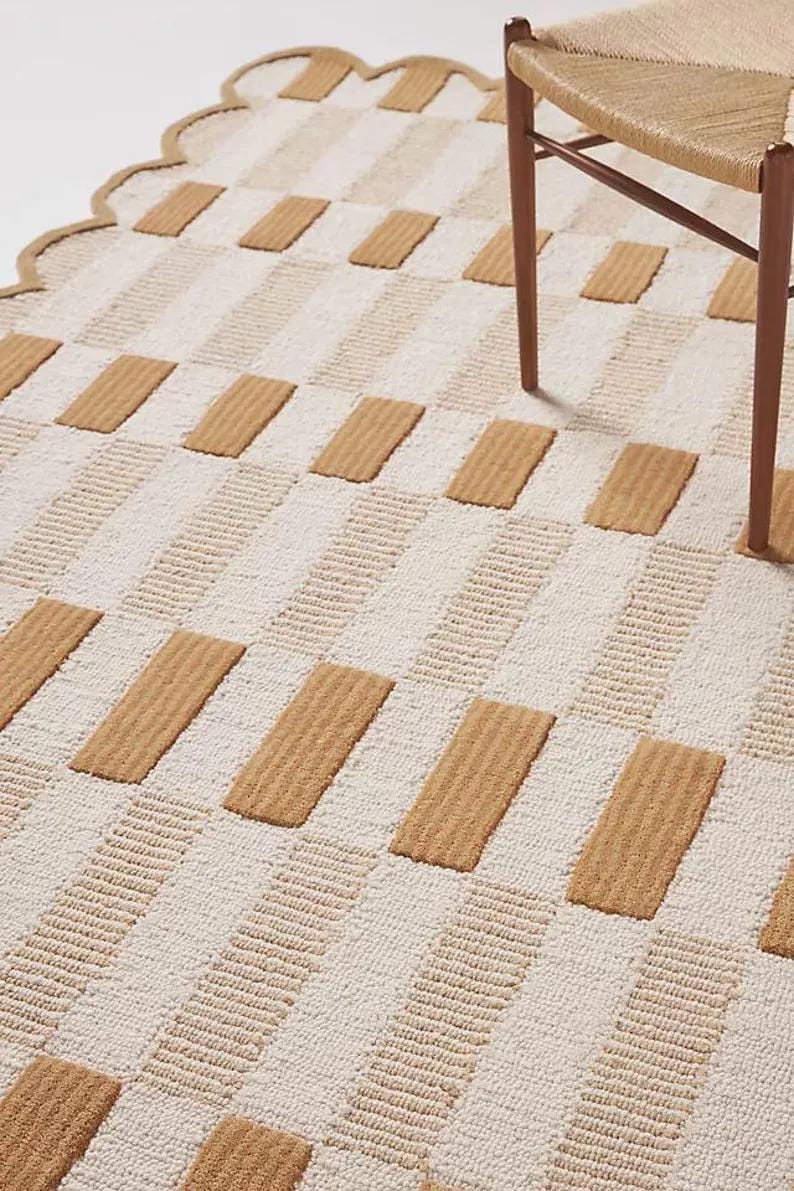 May include: A beige and white patterned rug with a scalloped edge. The rug features alternating rows of beige rectangular blocks and white striped sections. A wooden chair leg is visible.