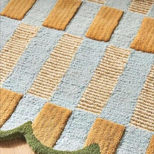 May include: A rectangular rug with a geometric pattern. The rug features alternating sections of light blue, tan, and mustard yellow. A green, wavy border trims the edge of the rug. The rug is made of a soft, textured material.