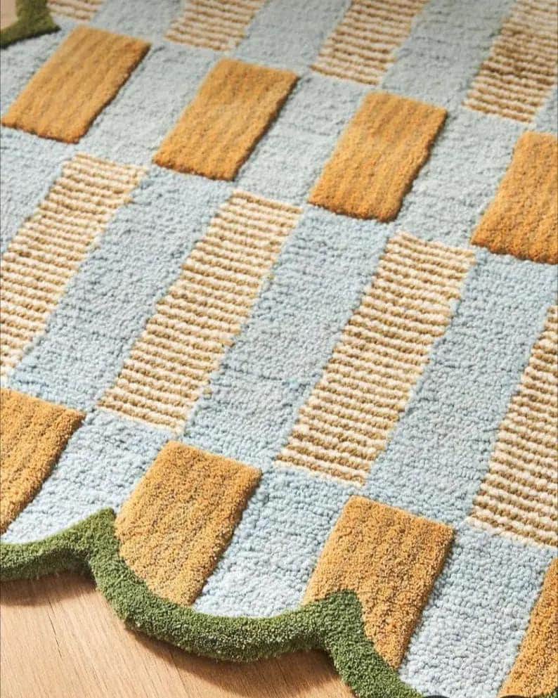 May include: A rectangular rug with a geometric pattern. The rug features alternating sections of light blue, tan, and mustard yellow. A green, wavy border trims the edge of the rug. The rug is made of a soft, textured material.