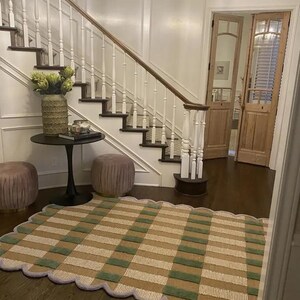 May include: A patterned rug with a scalloped edge, featuring green, beige and white stripes. The rug is placed on a dark wood floor in a home entryway, near a staircase and a small table.