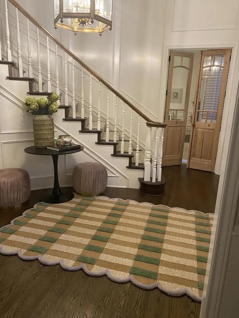 May include: A patterned rug with a scalloped edge, featuring green, beige and white stripes. The rug is placed on a dark wood floor in a home entryway, near a staircase and a small table.