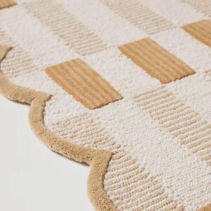 May include: A beige and white patterned rug with a scalloped edge. The rug features a repeating pattern of rectangular shapes in beige and white, creating a textured surface. The border is a solid beige colour.