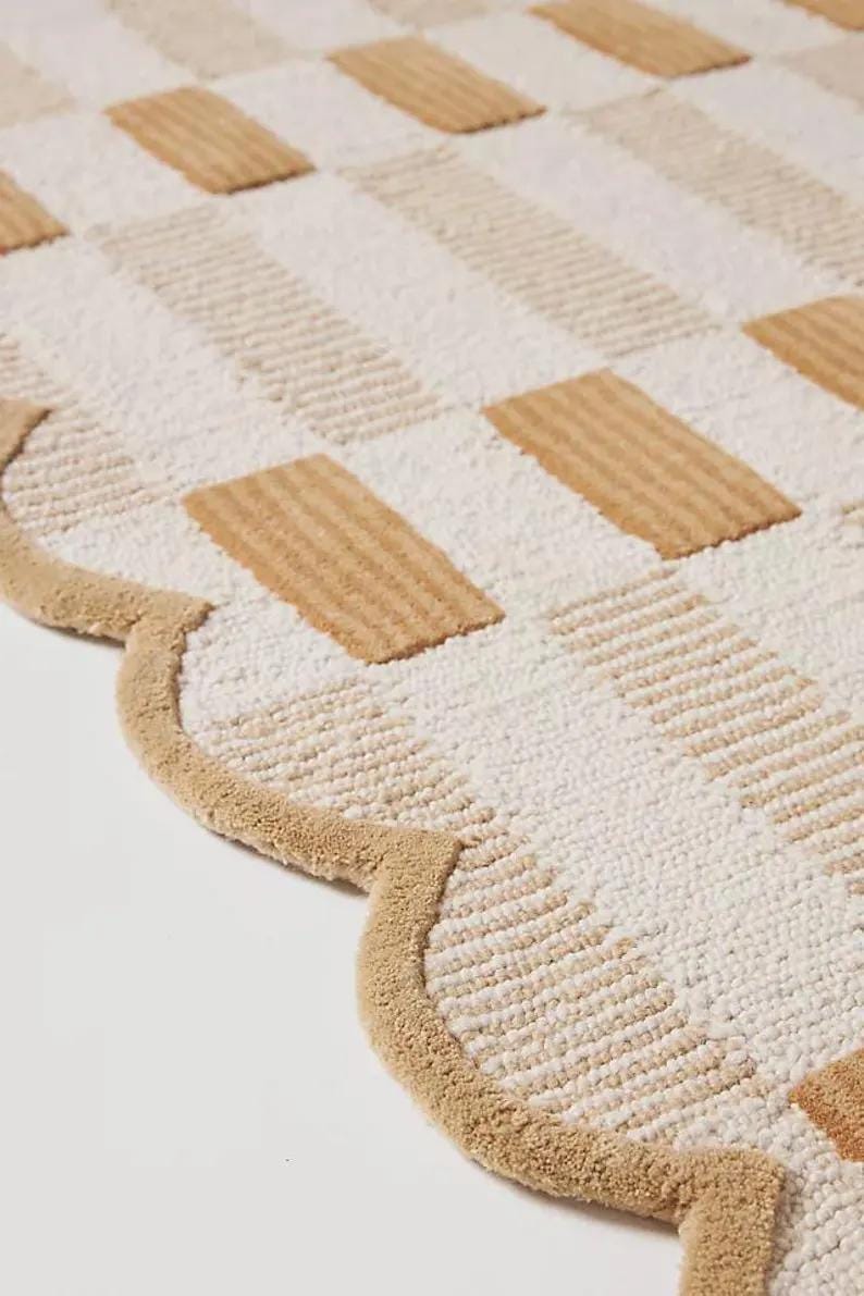 May include: A beige and white patterned rug with a scalloped edge. The rug features a repeating pattern of rectangular shapes in beige and white, creating a textured surface. The border is a solid beige colour.