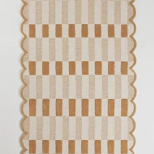 May include: Rectangular rug with a tan and cream checkered pattern. The rug has a scalloped edge in a tan colour. The pattern consists of vertical stripes of tan and cream, creating a grid-like design. The rug is made of a soft, textured material.