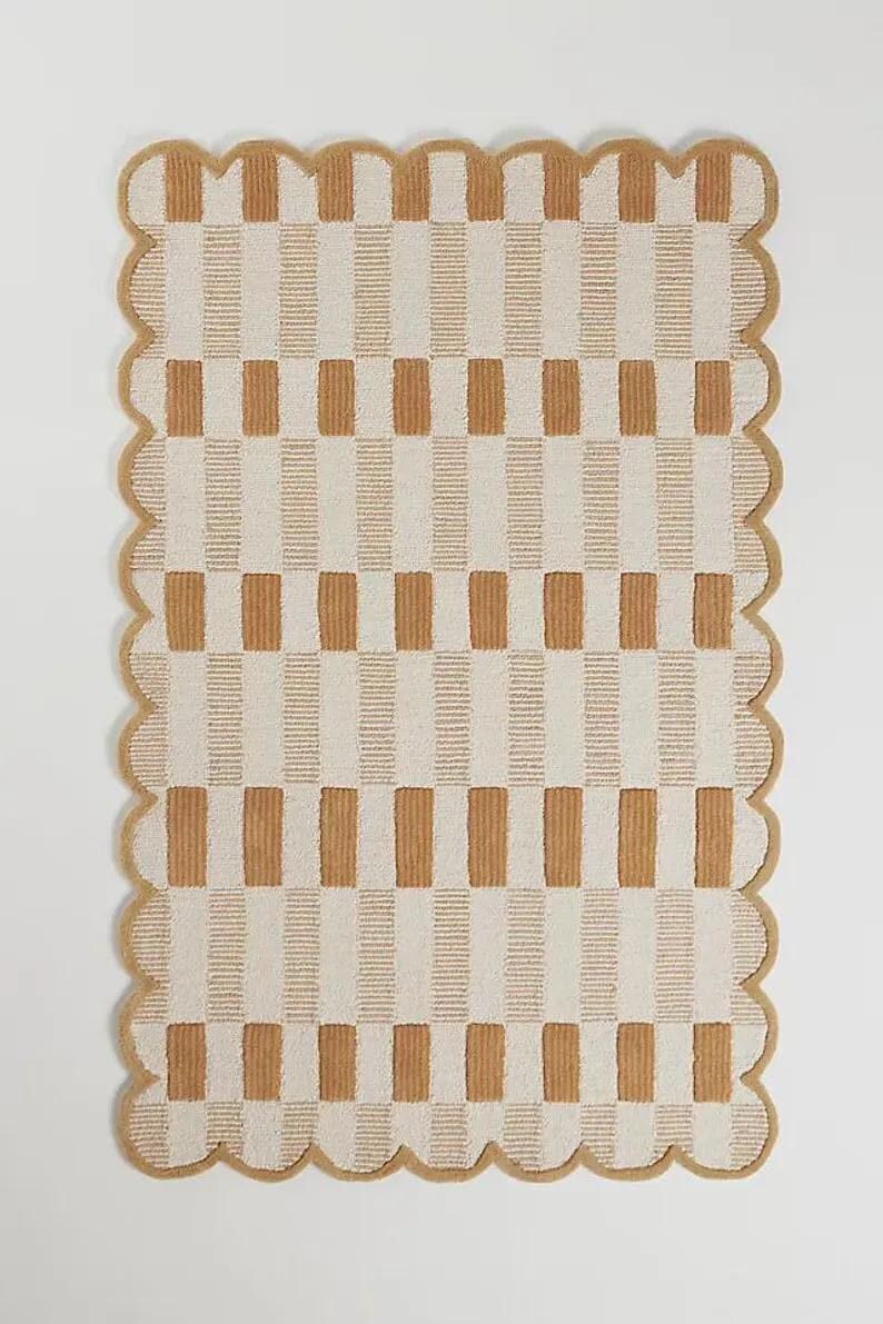 May include: Rectangular rug with a tan and cream checkered pattern. The rug has a scalloped edge in a tan colour. The pattern consists of vertical stripes of tan and cream, creating a grid-like design. The rug is made of a soft, textured material.