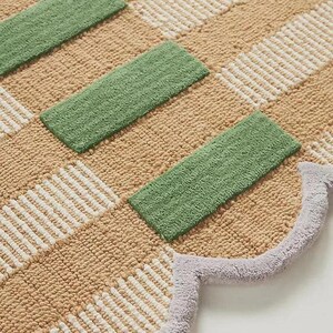 May include: A patterned rug with a geometric design. The rug has alternating sections of beige, white, and green rectangular shapes. A light purple scalloped edge borders the rug.