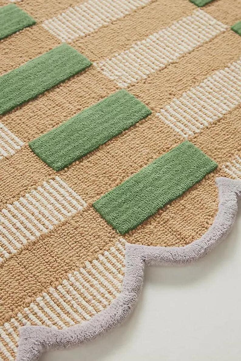 May include: A patterned rug with a geometric design. The rug has alternating sections of beige, white, and green rectangular shapes. A light purple scalloped edge borders the rug.