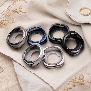 May include: A selection of watch cases in shades of silver, black, and blue. The cases feature a contemporary design with a transparent centre and coloured highlights. A gold ring is placed on a small white dish.