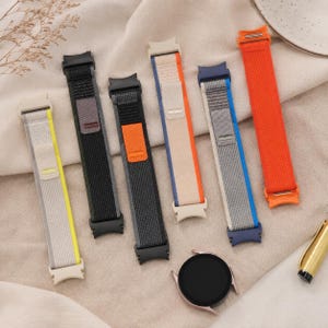 May include: An assortment of colorful watch bands in various shades, including gray, black, orange, and blue. The bands are made of a woven fabric and are displayed with a rose gold smartwatch. A gold pen is also visible.