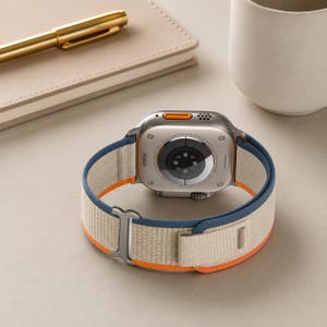 May include: A silver smartwatch with a beige, blue, and orange band. The watch is lying on a light-colored surface, with a pen, notebook, and mug in the background. The watch band is made of a woven material.