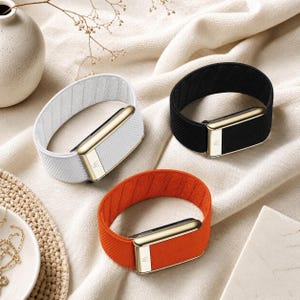 May include: Three fitness trackers in white, black, and orange. Each tracker has a gold-colored rectangular center piece with a textured surface and a gold-colored clasp. The bands are made of a woven material.