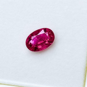 May include: A vibrant, oval-cut gemstone with a deep, saturated magenta-red hue. The stone features a faceted surface that reflects light, creating a brilliant, sparkling appearance across its polished, symmetrical, and translucent crystalline structure.