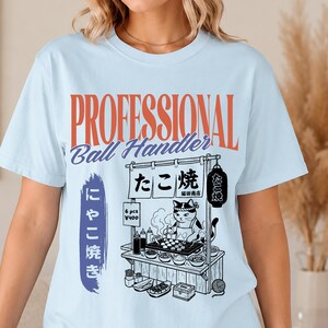 Puede incluir: Una camiseta azul claro con el texto "PROFESSIONAL Ball Handler" en naranja y azul. Debajo, un gráfico en blanco y negro muestra a un gato cocinando takoyaki en un puesto de comida con texto japonés y una linterna.