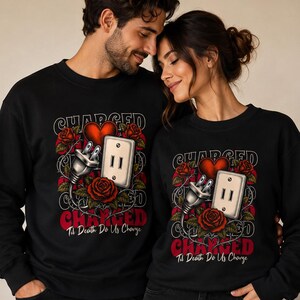 Peut inclure: Deux sweat-shirts noirs à col rond avec une impression graphique représentant un cœur rouge, une prise électrique et une prise murale entourées de roses rouges. Le texte indique "CHARGED" en grandes lettres et "Til Death Do Us Charge" en dessous.