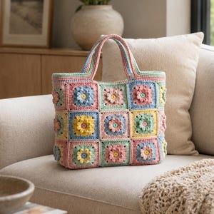 Op de afbeelding: Een handgemaakte gehaakte tas opgebouwd uit meerdere vierkante panelen met verhoogde bloemmotieven in pasteltinten zoals roze, geel, blauw en groen. De tas is voorzien van twee bijpassende gehaakte handvatten en een gestructureerde rechthoekige bodem.