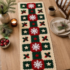 May include: A rectangular crochet table runner featuring a repeating pattern of cream, dark green, and red squares. The design includes white snowflake motifs, small beige stars, and red berry clusters. The runner is crafted with a textured yarn finish.