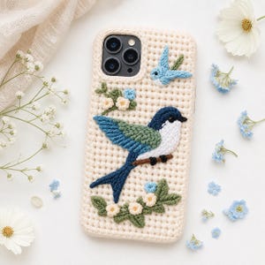 May include: Cream-colored crochet phone case featuring a detailed, colorful bird design. The bird has blue, green, and white accents, perched on a brown branch with floral embellishments. A smaller bird and flowers are also present.