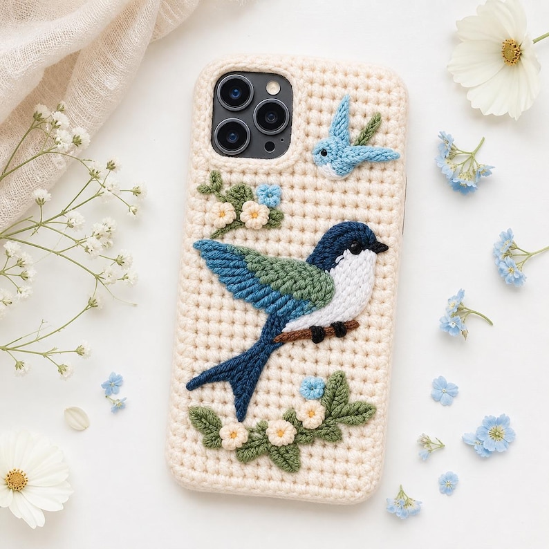 May include: Cream-colored crochet phone case featuring a detailed, colorful bird design. The bird has blue, green, and white accents, perched on a brown branch with floral embellishments. A smaller bird and flowers are also present.
