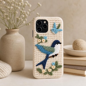 May include: Cream-colored crochet phone case featuring a blue, green, and white bird perched on a branch. The case has a textured appearance with a small blue bird and floral accents. The phone case is designed to fit a smartphone.