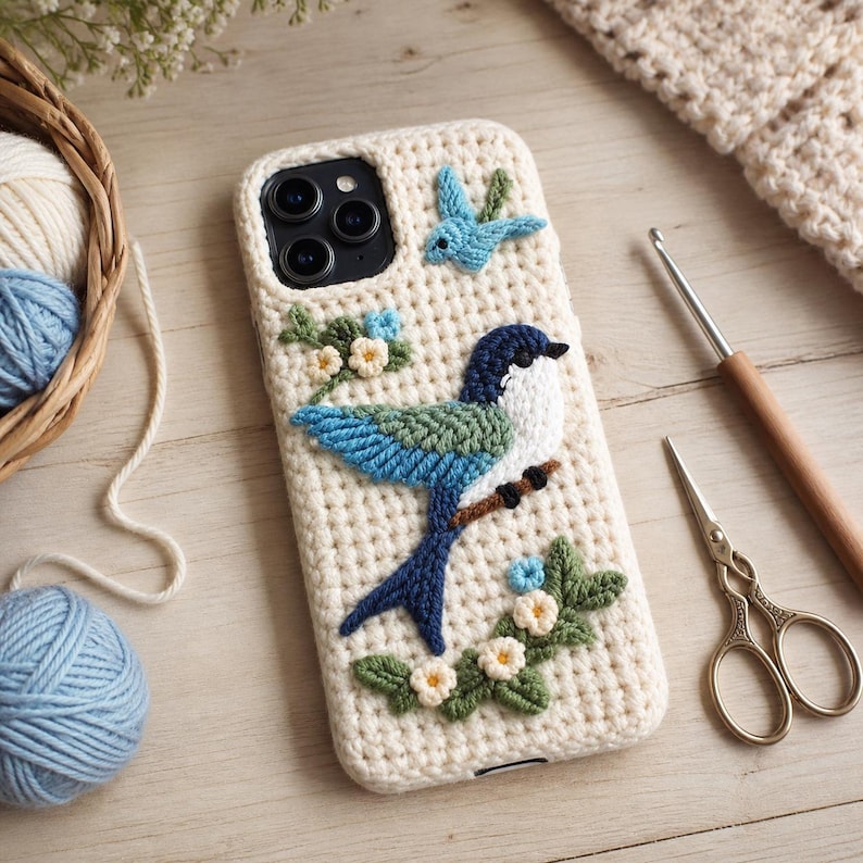 May include: Cream-colored phone case with a crocheted bird design. The bird features blue and green wings, a white chest, and a dark blue head. Additional crocheted flowers and a small bird are also present. A basket of yarn and crochet tools are in the background.