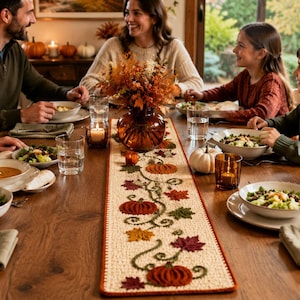 Pode incluir: Um caminho de mesa retangular de cor creme com um design bordado e texturizado de abóboras cor de laranja, vinhas verdes e folhas de outono multicoloridas em tons de vermelho, laranja e amarelo, com uma borda costurada em laranja escuro.