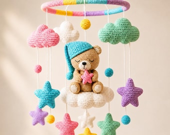 May include: A crochet baby mobile featuring a central brown teddy bear wearing a blue nightcap and holding a pink star while sitting on a white cloud. The mobile includes colorful hanging clouds, stars, and spheres suspended from a striped ring.
