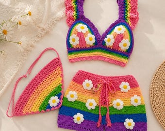Könnte beinhalten: Ein Häkelset bestehend aus einem Bralette, einer Shorts und einem dreieckigen Kopftuch. Jedes Teil zeigt ein lebendiges Regenbogen-Streifenmuster mit aufgesetzten weißen Gänseblümchen-Applikationen. Das Bralette hat Rüschenträger und die Shorts einen Kordelzug.