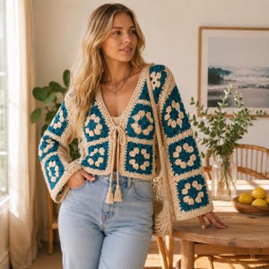 May include: A long-sleeved crochet cardigan featuring a granny square pattern in teal and cream. The garment has a V-neckline, wide sleeves, and a front tie closure with two dangling tassels. The crochet construction creates a textured floral motif.