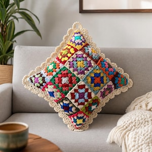 Könnte beinhalten: Ein quadratischer Kissenbezug aus Häkelarbeit mit einem lebendigen Granny-Square-Muster in verschiedenen Farben wie Rot, Blau, Gelb, Grün und Lila. Das Kissen ist mit einem cremefarbenen, wellenförmigen Häkelrand und einer kleinen Aufhängeschlaufe versehen.