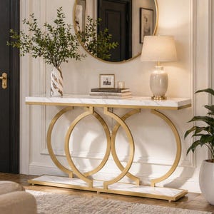 Op de afbeelding: Een console met een marmeren blad en een gouden metalen frame. De tafel heeft drie grote, in elkaar grijpende gouden ringen als steunen. Een vaas met groen, een lamp en boeken worden op de tafel tentoongesteld. De kamer heeft een spiegel en een witte muur.
